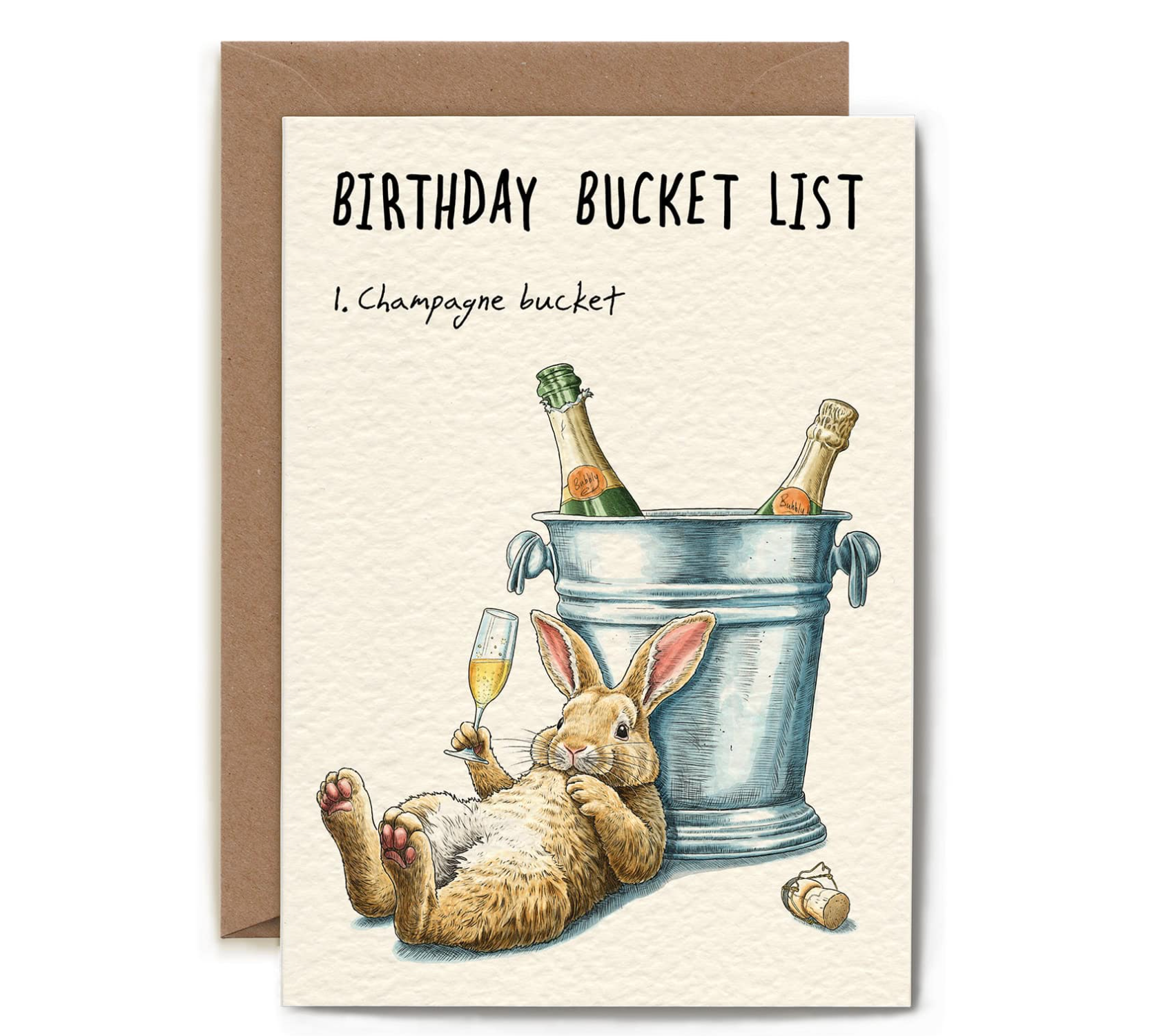 A greeting card with a brown envelope behind it, featuring the text 'BIRTHDAY BUCKET LIST' and '1. Champagne bucket' above an illustration of a rabbit lying on its back beside a metal bucket with two champagne bottles and a glass of champagne.
