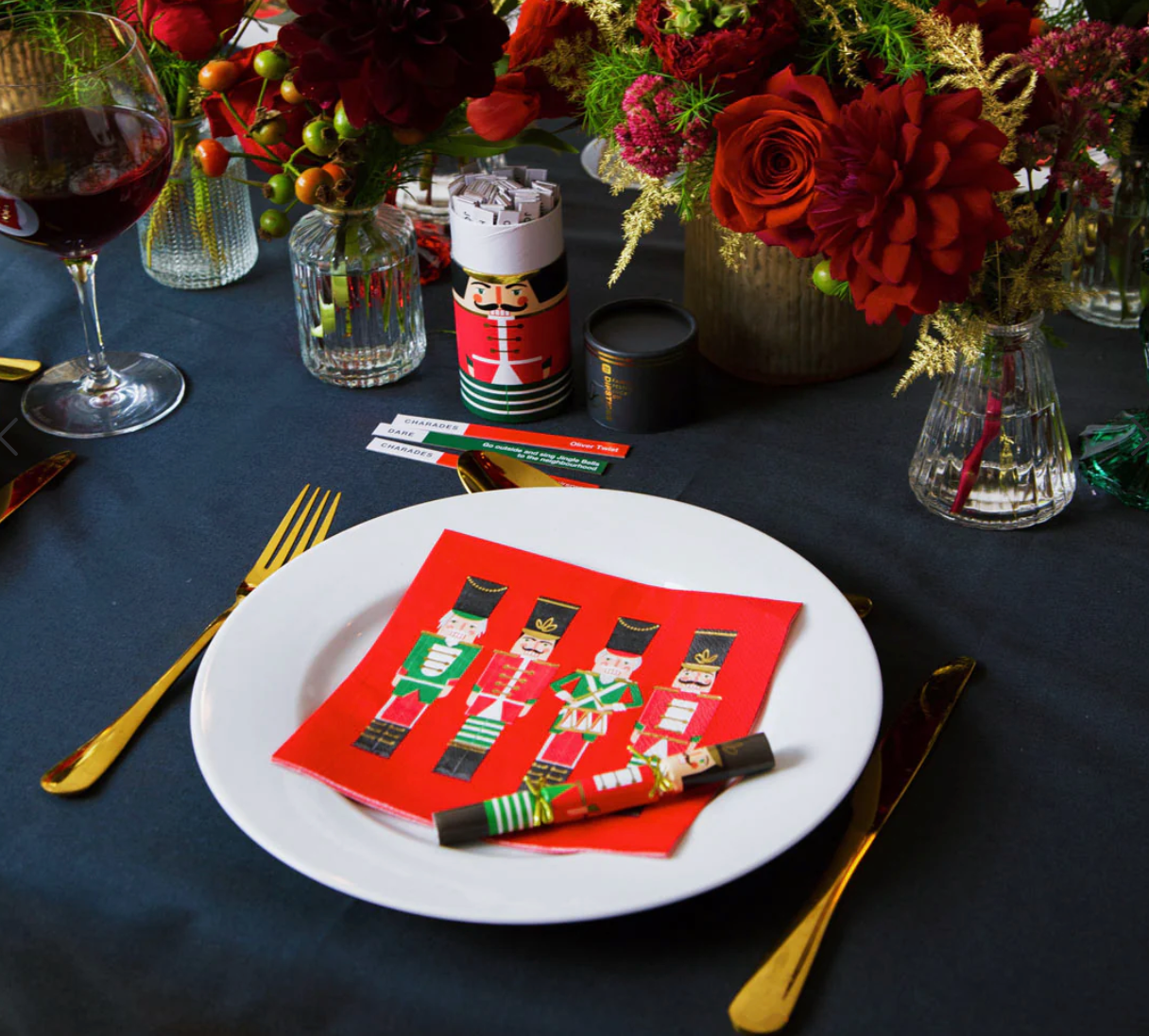 Table setting with a white plate, red napkin with nutcracker design, and wine glass on a dark tablecloth.