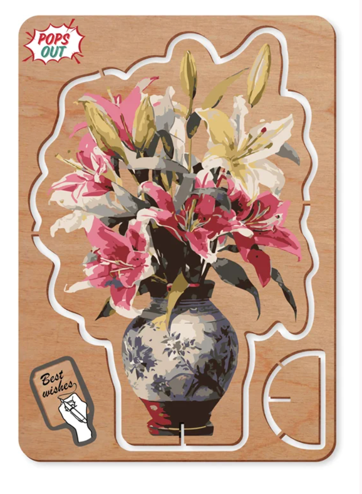 A wooden 'Pops Out' card featuring a floral design of pink and white lilies in a blue and white patterned vase, set against a light wood background with white puzzle piece outlines.