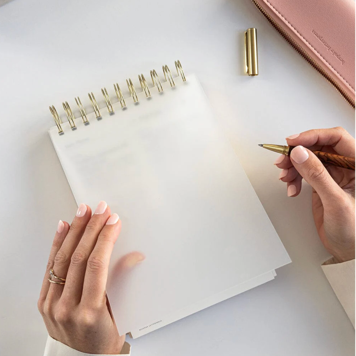 Person holding a pen over an open notebook with a pink notebook in the background