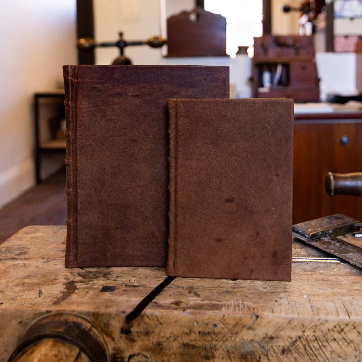 Two sizes of SB Libris Classic Full Leather Journals in aged goatskin, showing their warm brown tones and subtle spine detailing on a traditional workbench.