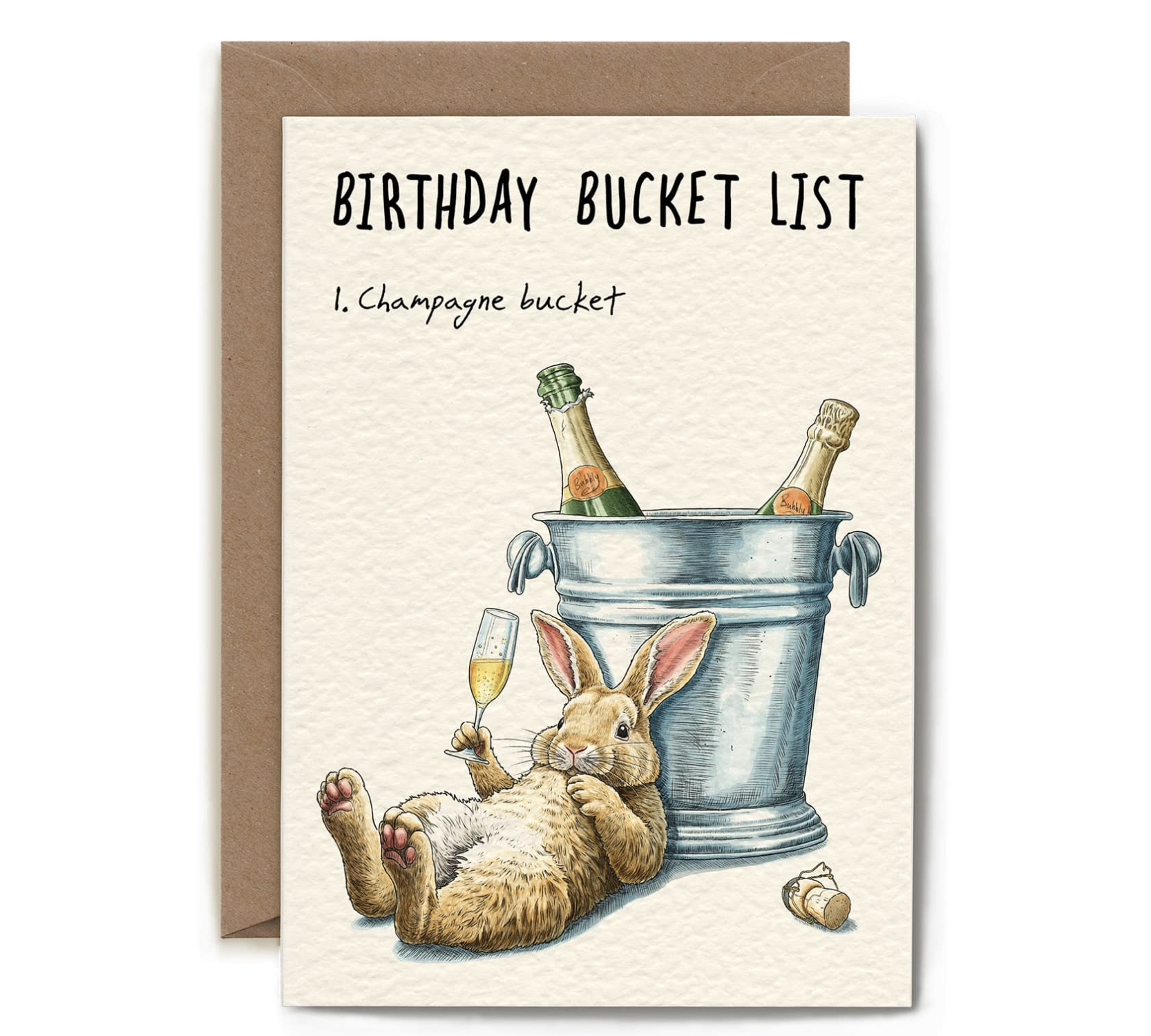 A greeting card with a brown envelope behind it, featuring the text 'BIRTHDAY BUCKET LIST' and '1. Champagne bucket' above an illustration of a rabbit lying on its back beside a metal bucket with two champagne bottles and a glass of champagne.