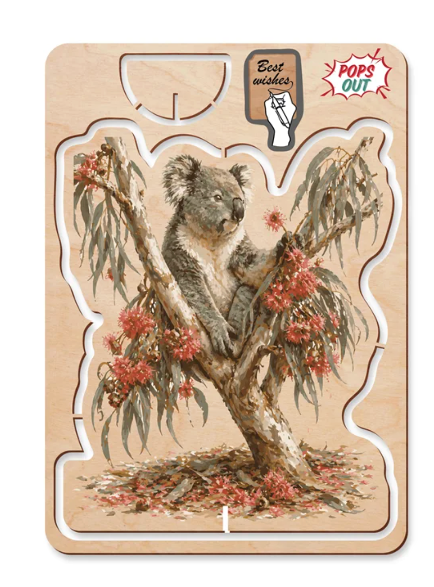 A wooden pop up card featuring an illustration of a koala sitting in a eucalyptus tree with red flowers. The puzzle is displayed on its wooden backing card, which includes a 'Best wishes' stamp and a 'POPS OUT' logo.