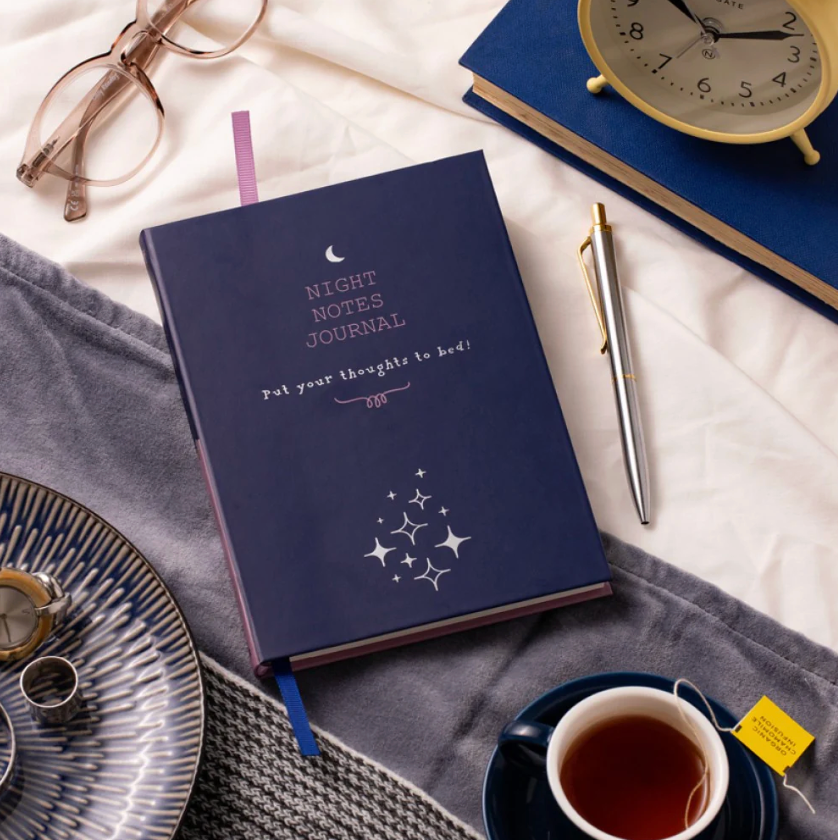 Navy blue 'Night Notes Journal' on a white surface with a cup of tea, glasses, and alarm clock.