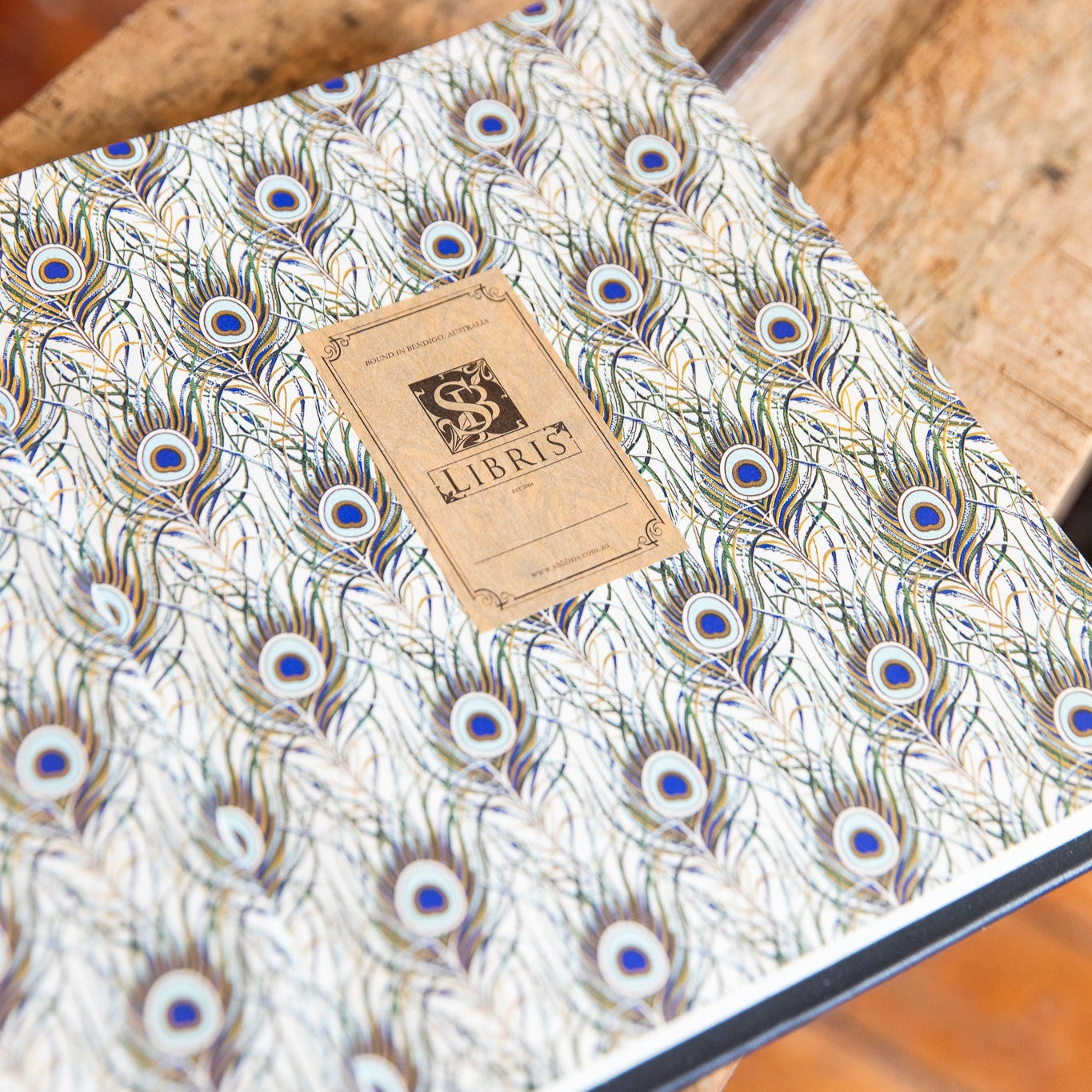 Notebook with peacock feather pattern and Libris brand tag on a wooden surface