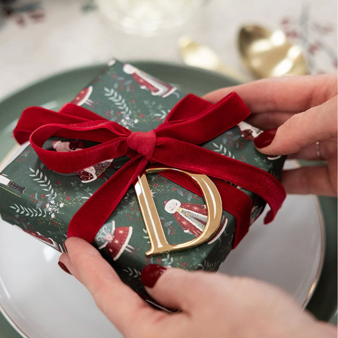 Person holding a gift wrapped in green paper with a red ribbon and gold letter 'D'.