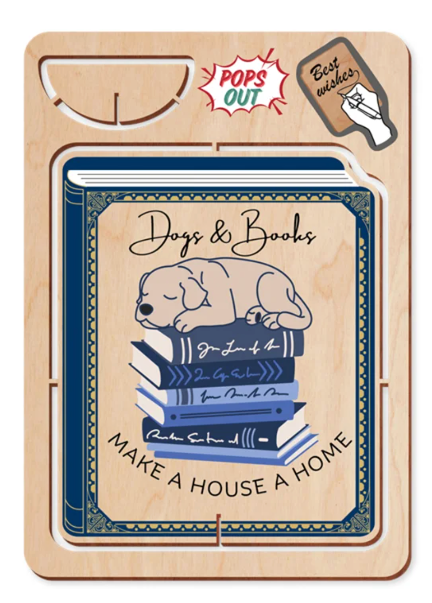 A wooden gift card or book-shaped card with a printed illustration of a sleeping dog on a stack of books and the text 'Dogs & Books' and 'MAKE A HOUSE A HOME.' The card is mounted on a light wood backing with a die-cut hole and includes a 'Pops Out' burst and a 'Best wishes' stamp graphic near the top.