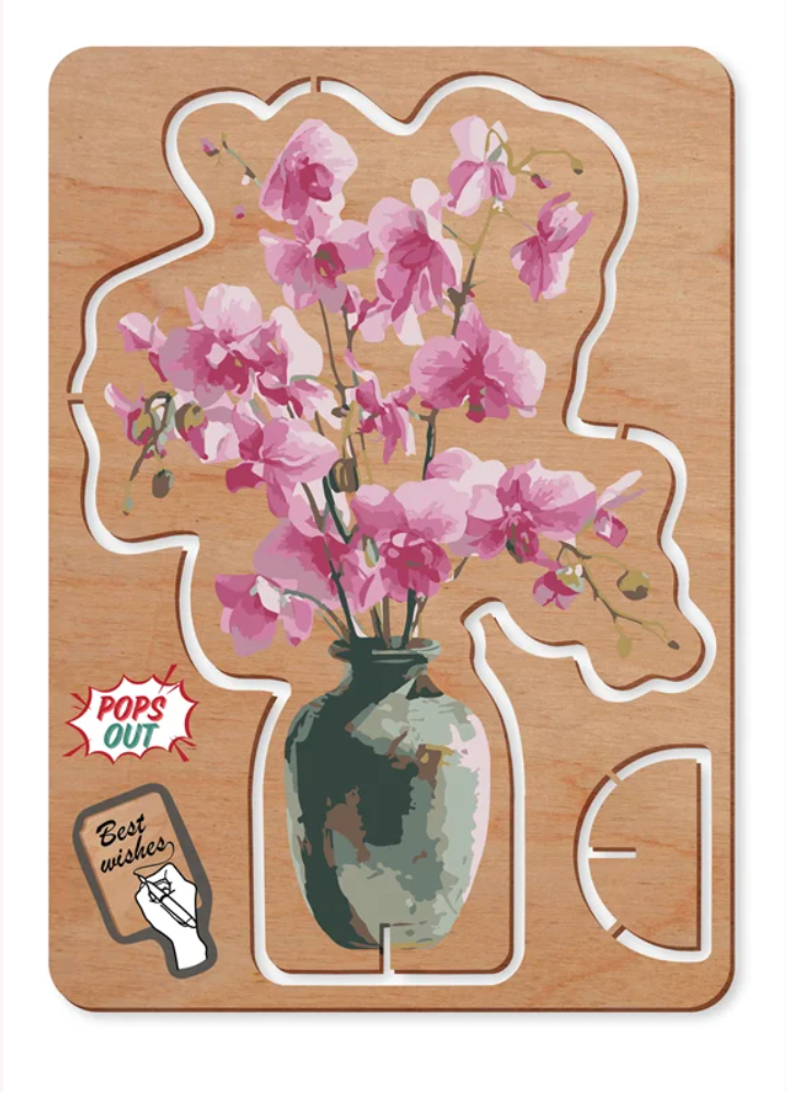A wooden Pop Up cardfeaturing a pink orchid bouquet in a vase, shown assembled on a light wood background with cut-out flower and vase shapes. Decorative text badges read 'POPS OUT' and 'Best wishes'.